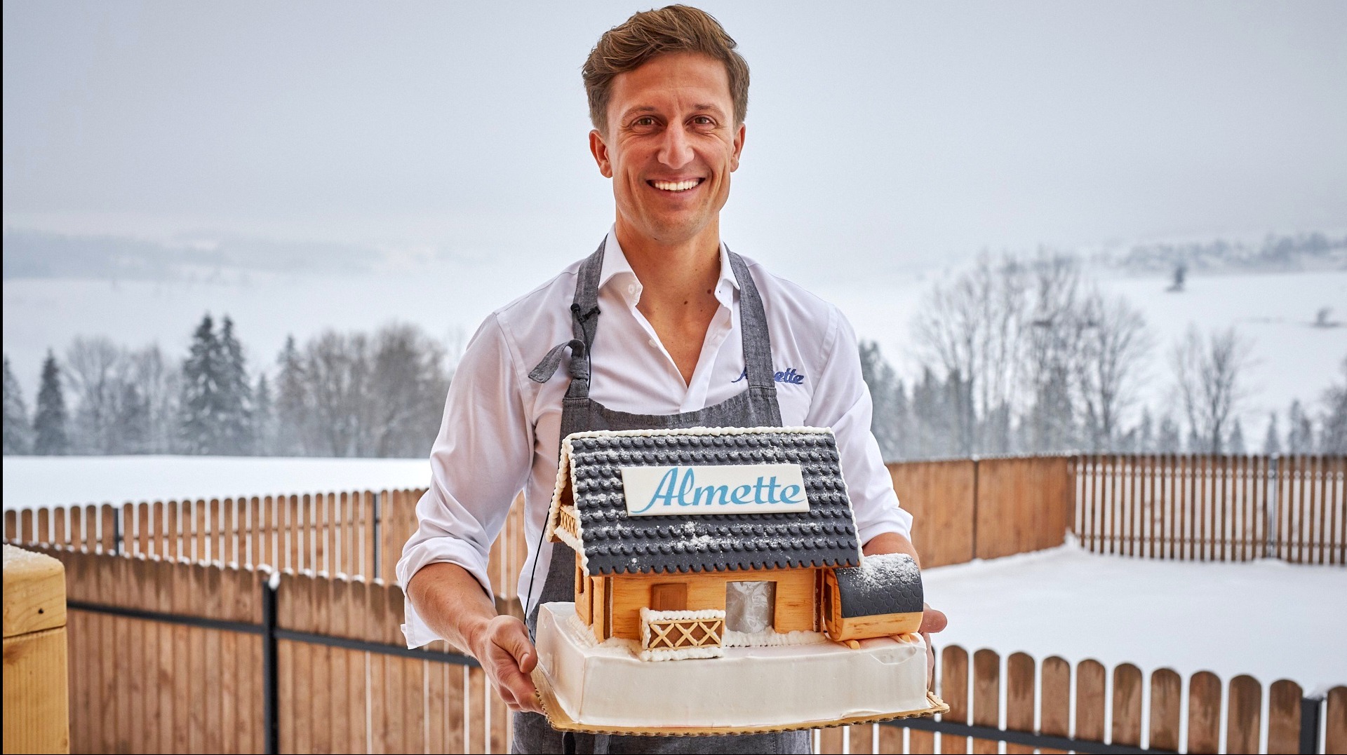 Uśmiechnięty mężczyzna w fartuchu prezentuje tort w kształcie drewnianego domku z napisem Almette na dachu, na tle zimowego krajobrazu z zalesionymi wzgórzami.