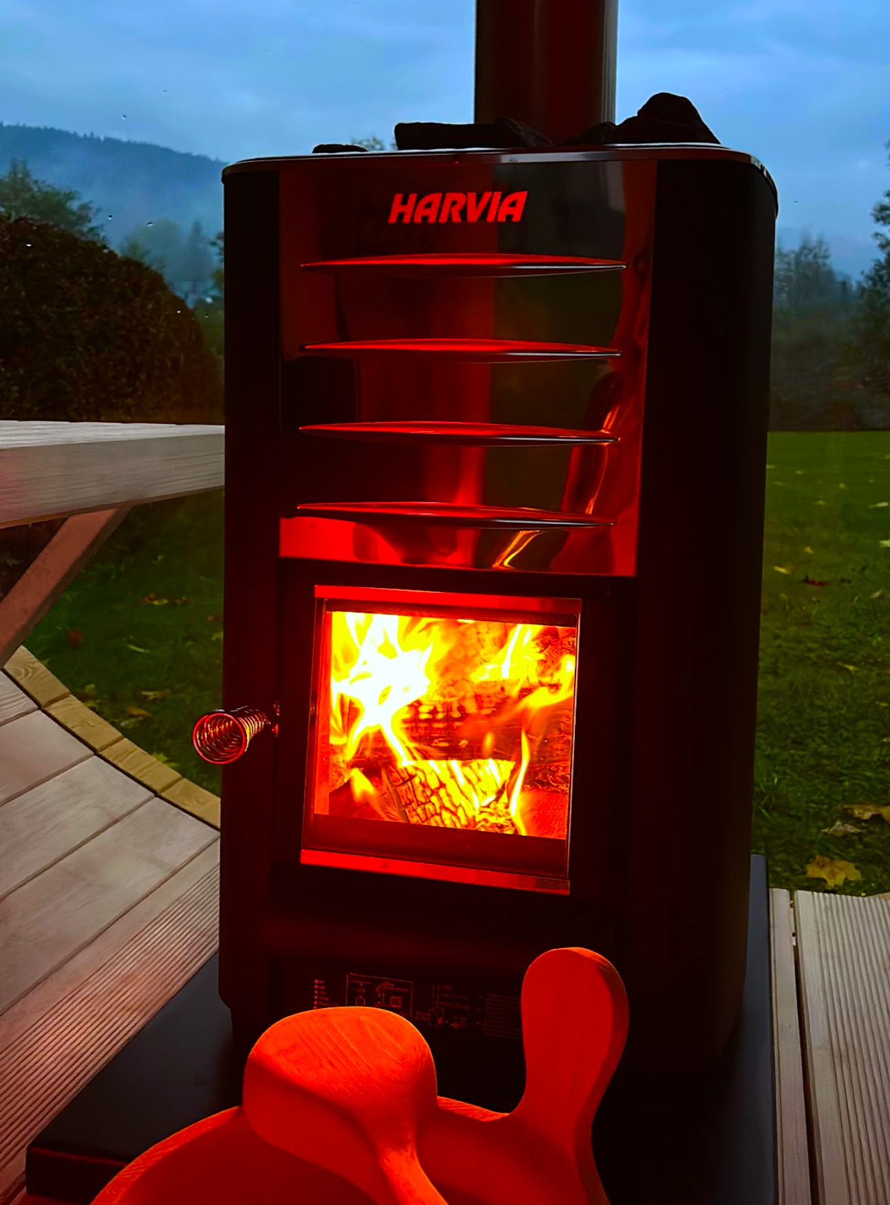 Opalana drewnem piec do sauny Harvia z płomieniami widocznymi przez szklane drzwiczki, obok drewniane akcesoria do sauny i widok na zewnątrz z zalesionym wzgórzem w tle.
