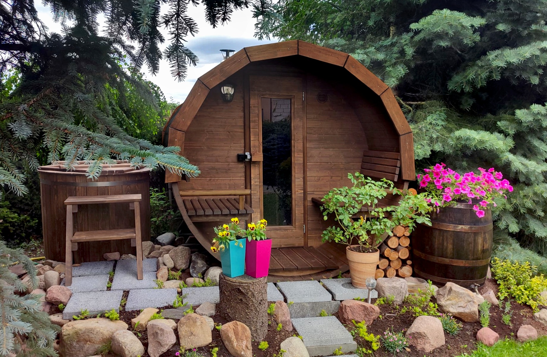 Drewniana beczka sauny ogrodowej z oknem i lampą, otoczona zielenią, kamieniami i kwiatami w kolorowych doniczkach, tworząca relaksującą przestrzeń.