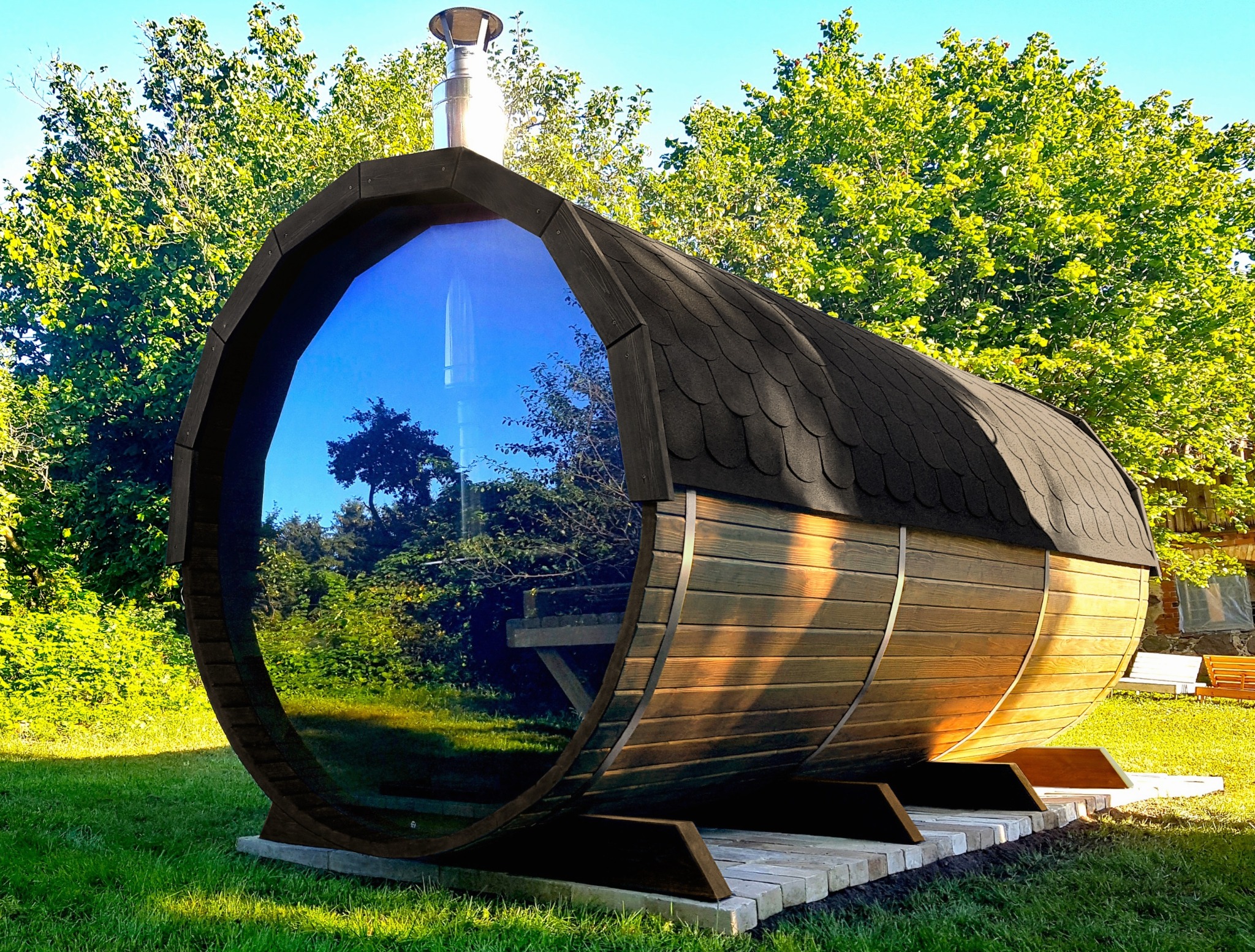 Drewniana sauna w kształcie beczki z panoramicznym oknem odbijającym otaczającą zieleń ogrodu, umieszczona na betonowej podstawie.
