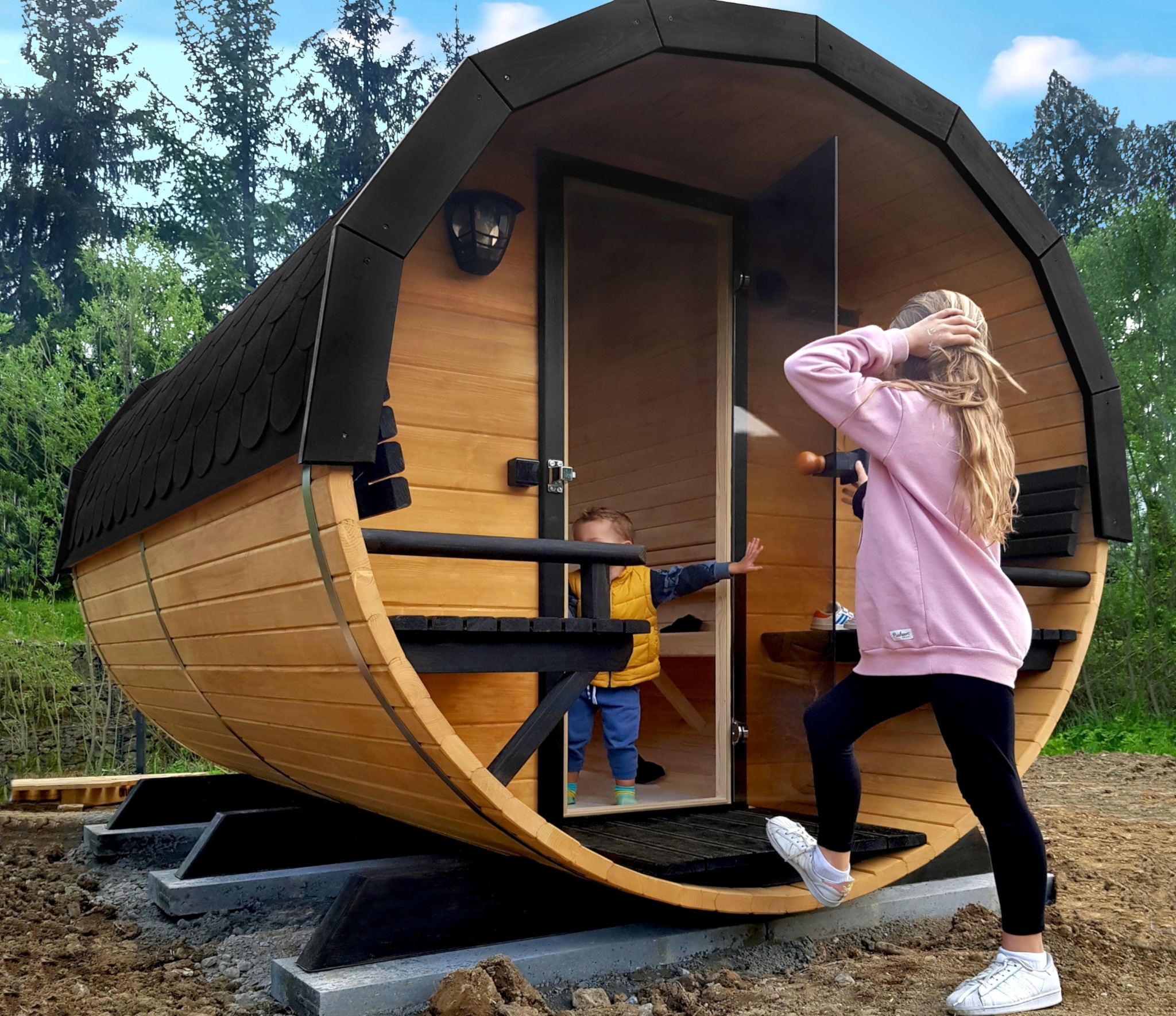 Drewniana sauna w kształcie beczki z czarnym dachem, zamontowana na betonowych fundamentach, z dziećmi w pobliżu wejścia.