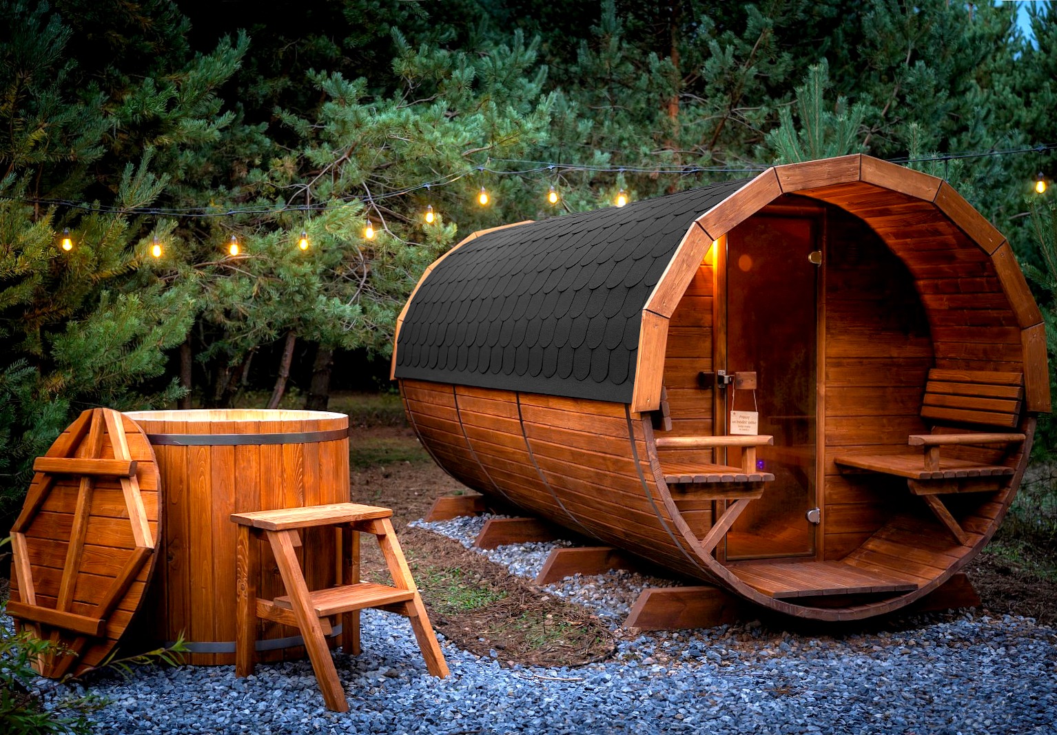 Drewniana beczka sauny z czarnym dachem z gontu bitumicznego, obok drewniana balia z pokrywą i schodkami, oświetlona girlandą żarówek, na tle zielonych drzew.