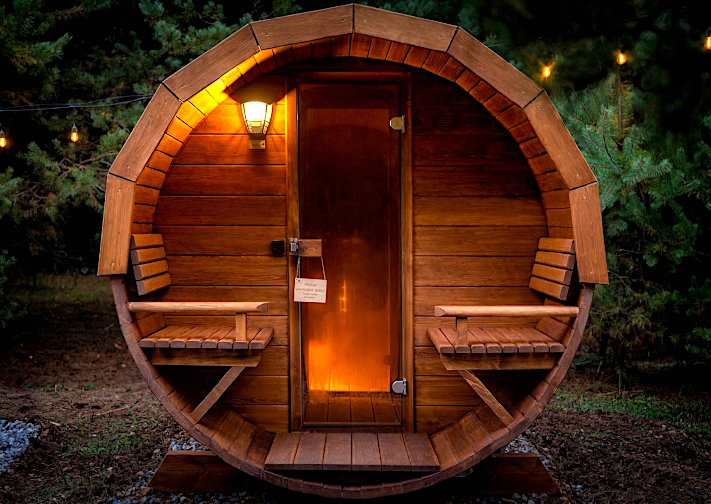 Drewniana beczka z sauną zewnętrzną z siedziskami po bokach, oświetlona lampą i girlandą, na tle zieleni.