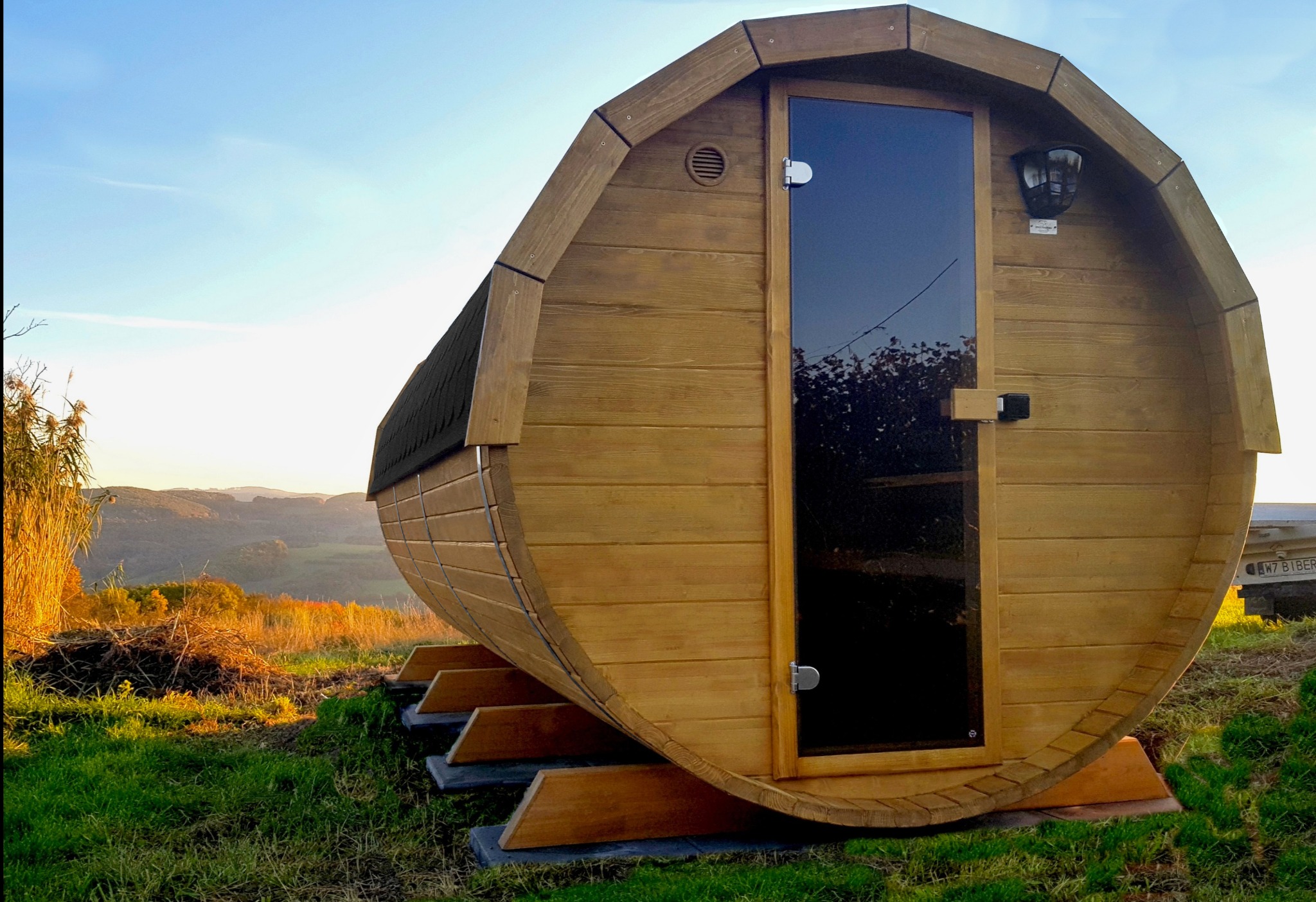Drewniana sauna w kształcie beczki z przeszklonymi drzwiami, umieszczona na zielonym wzgórzu z widokiem na dolinę w promieniach zachodzącego słońca.