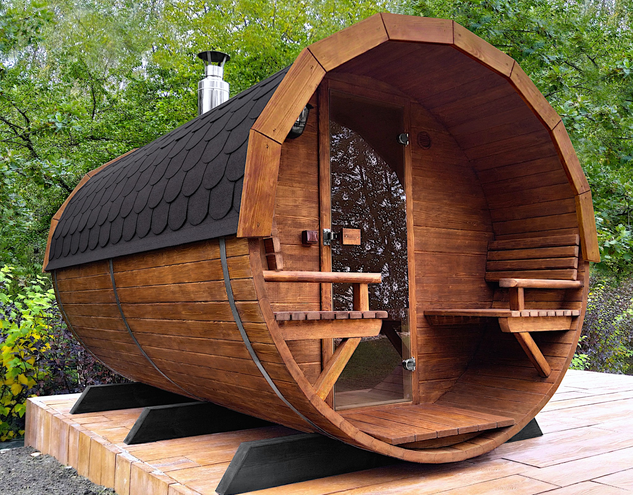 Drewniana sauna w kształcie beczki z czarnym, łuskowym dachem i szklanymi drzwiami, umieszczona na drewnianym podeście, na tle zieleni.