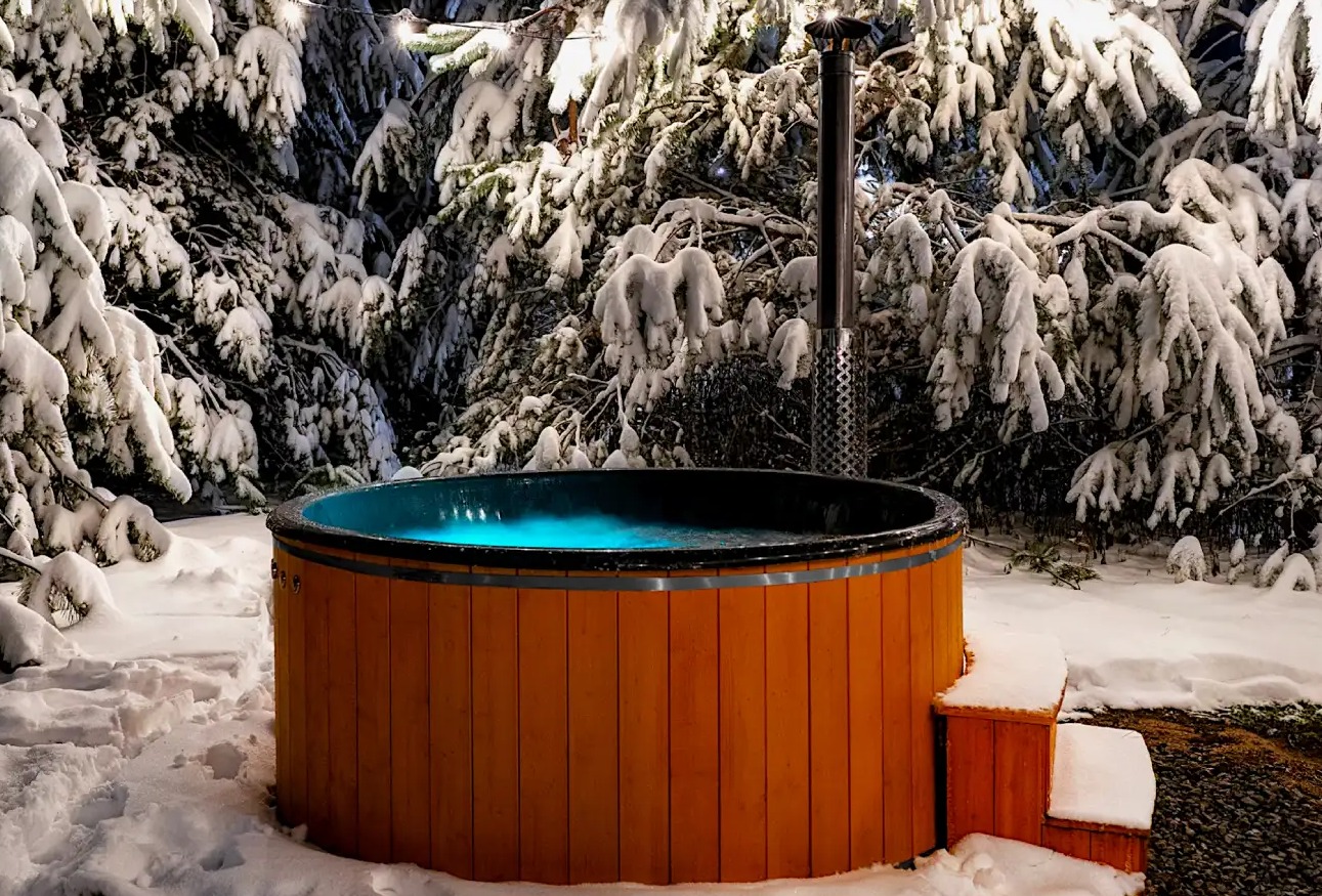 Drewniana balia ogrodowa z podświetlaną wodą, otoczona śniegiem i ośnieżonymi drzewami. Widoczne schodki wejściowe i komin. Zimowy krajobraz.