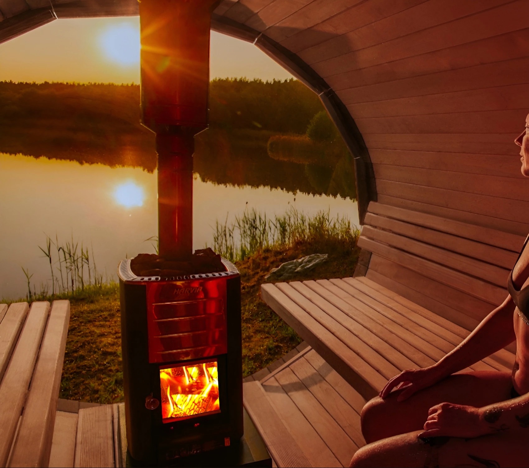 Oświetlona sauna beczka z widokiem na jezioro o zachodzie słońca. Wnętrze z drewnianymi ławkami i piecem Harvia. Kobieta siedzi na ławce.