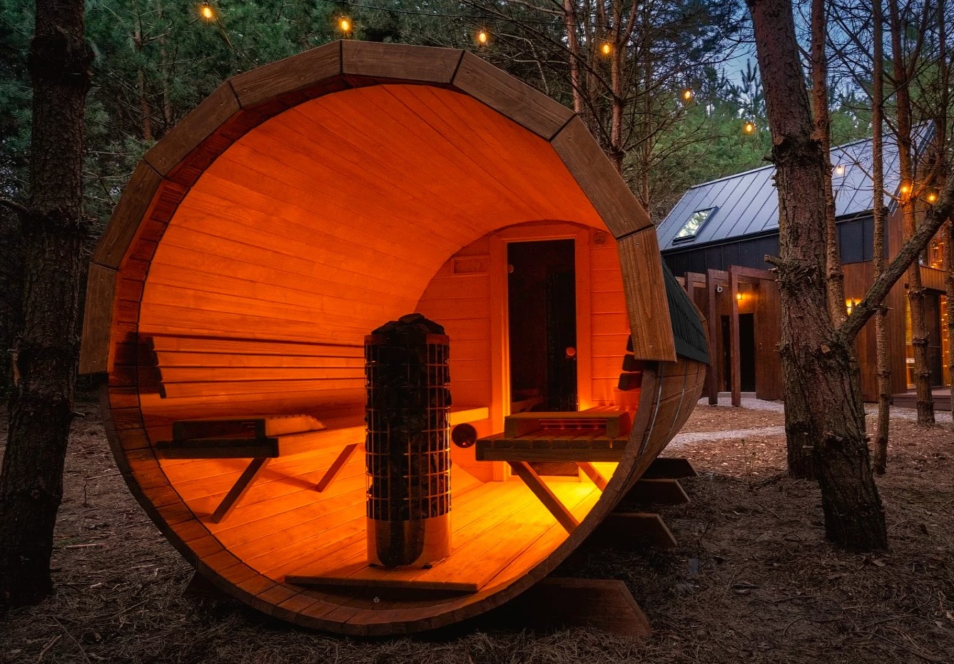 Drewniana sauna w kształcie beczki, oświetlona ciepłym, pomarańczowym światłem, stoi na zewnątrz w otoczeniu drzew. Widoczny piec i ławki wewnątrz sauny.