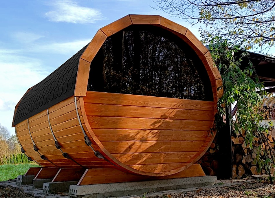 Drewniana beczka, prawdopodobnie sauna, z czarnym dachem, umieszczona na zewnątrz, na trawie, w słoneczny dzień. Widoczne drzewa w tle.
