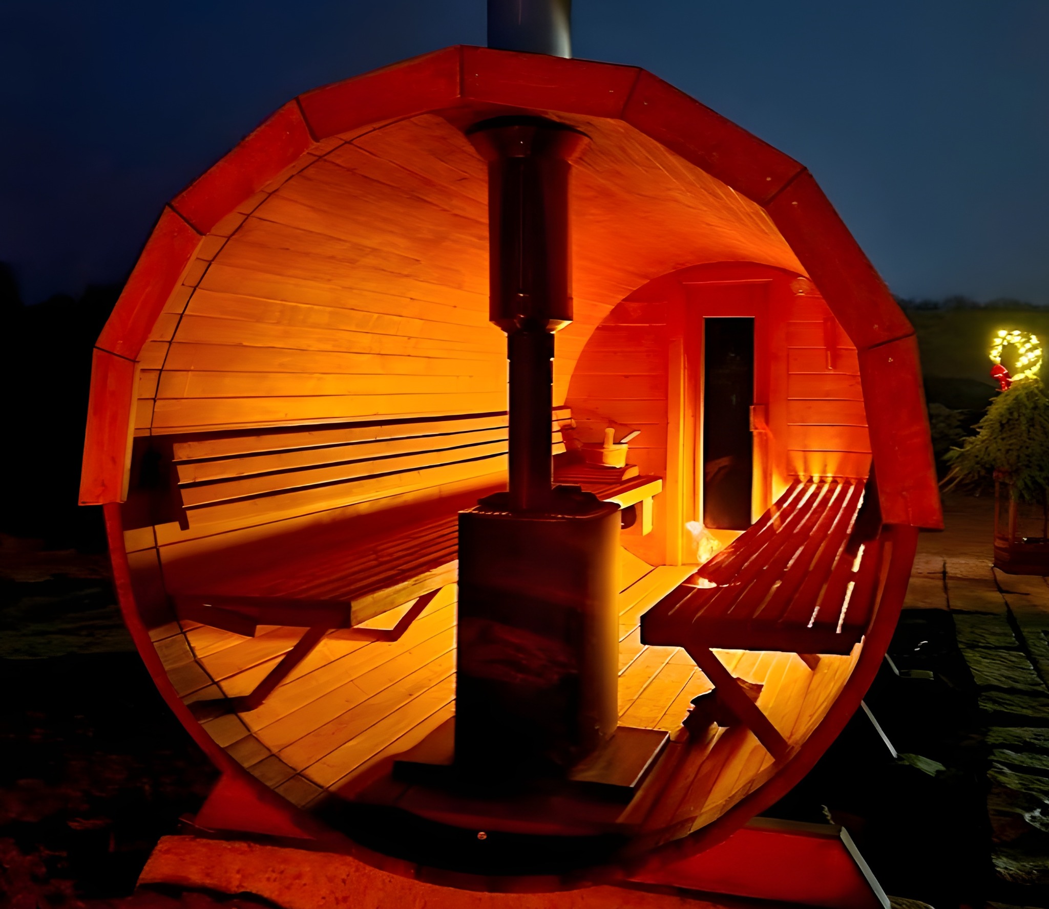 Beczkowa sauna z piecem w środku, oświetlona ciepłym światłem. Na zewnątrz ciemno, w tle widoczna choinka z lampkami. Unikalny kształt i atmosfera relaksu.