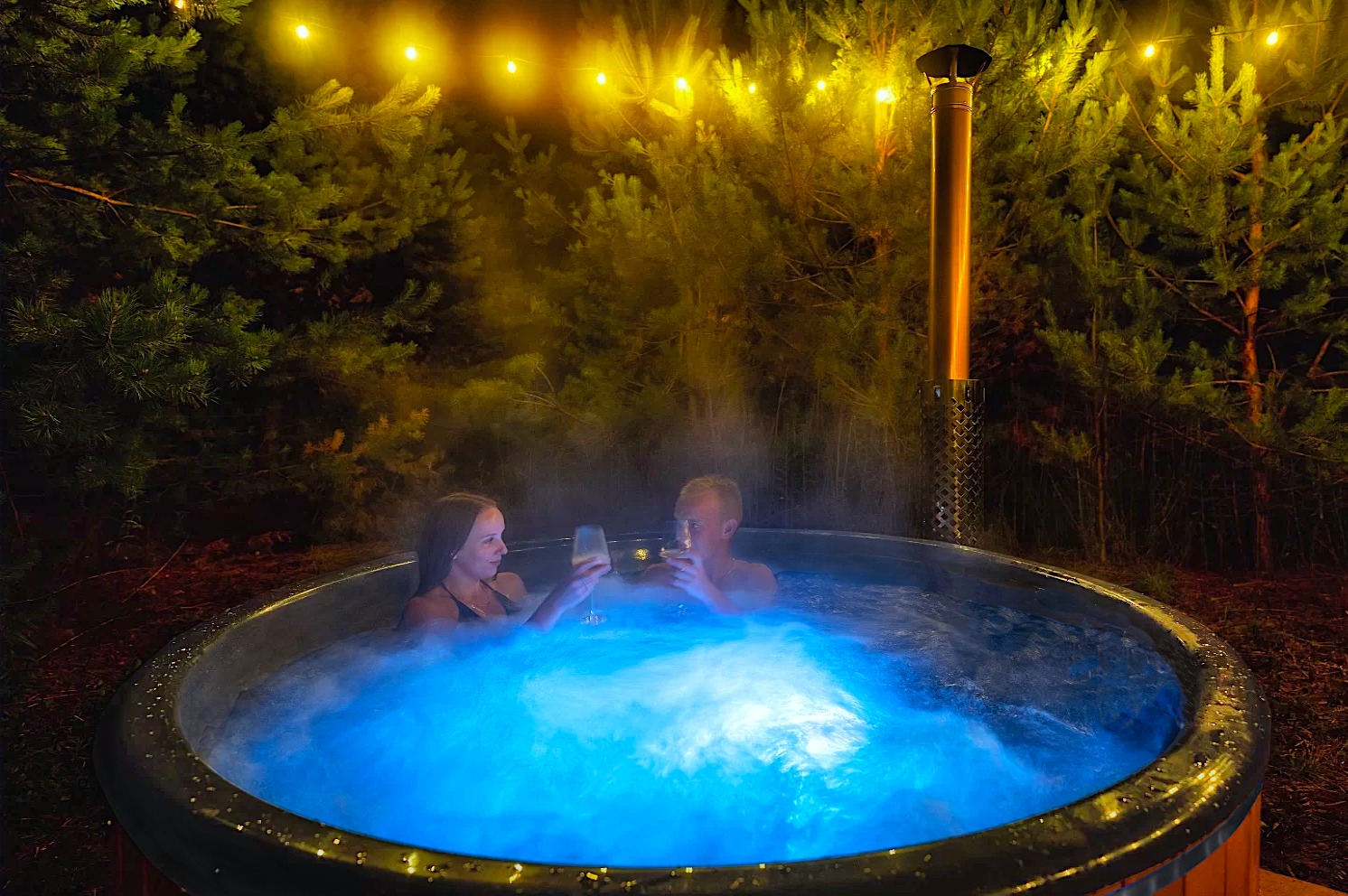 Para w balii ogrodowej z hydromasażem, podświetlonej na niebiesko, w otoczeniu iglaków i girlandy światełek, trzymająca kieliszki z winem.