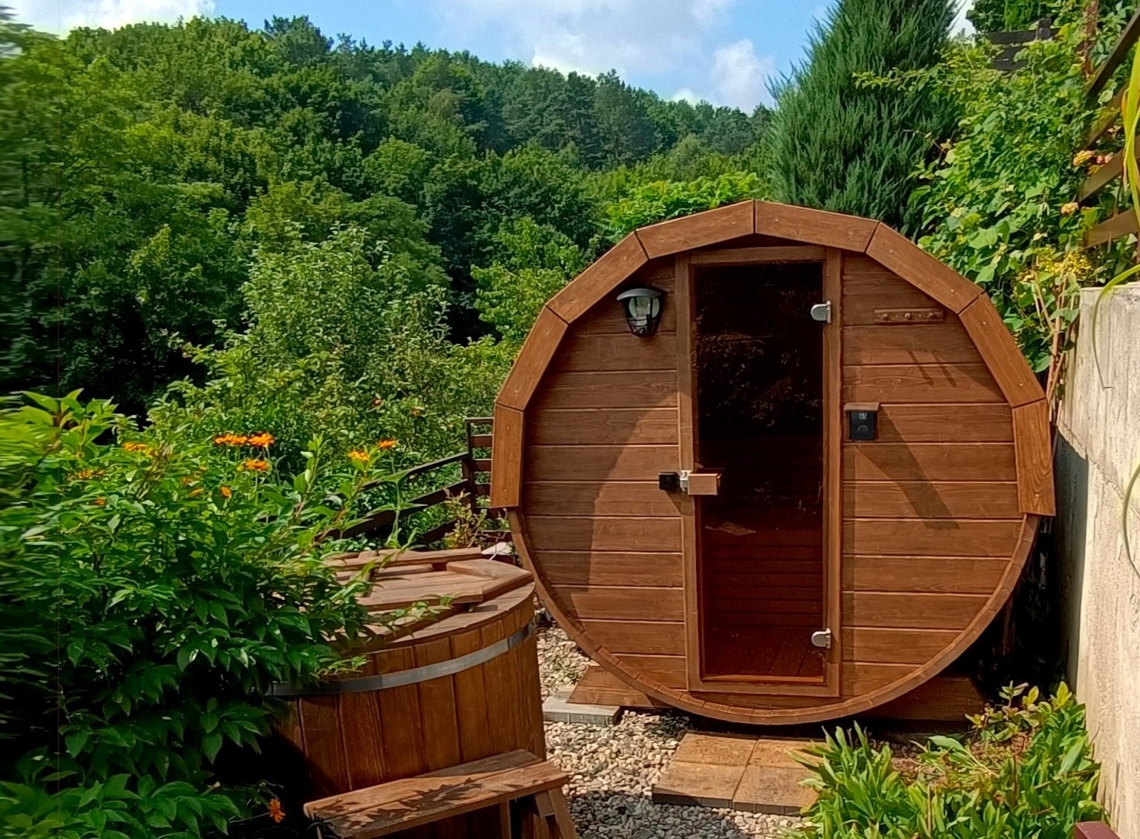 Drewniana beczka sauny z przeszklonymi drzwiami i lampą na zewnątrz, otoczona zielenią ogrodu. Obok stoi drewniana balia.
