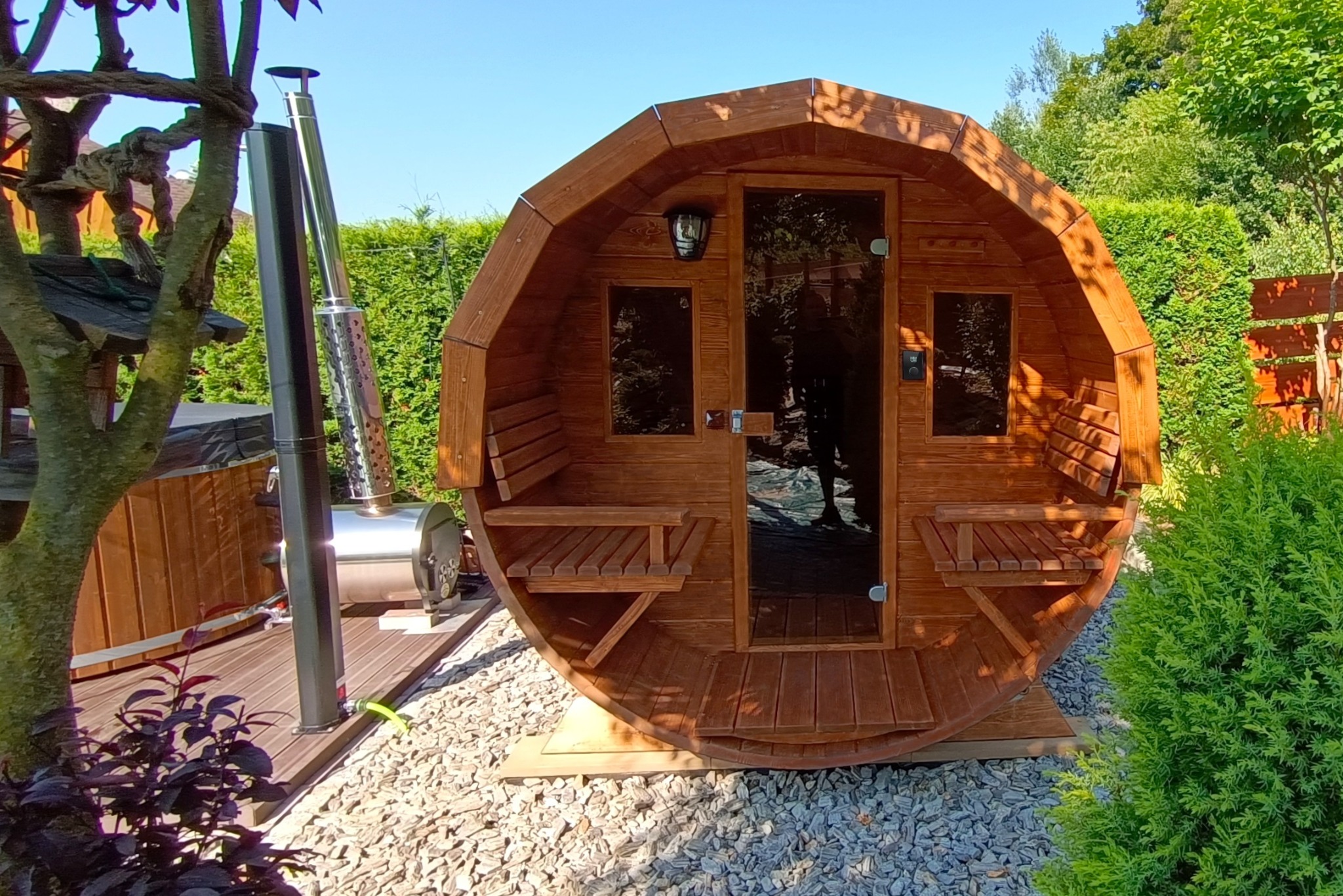 Drewniana sauna w kształcie beczki z piecem na zewnątrz, otoczona zielenią. Uwagę przyciąga nietypowy kształt sauny i jej umiejscowienie w ogrodzie.