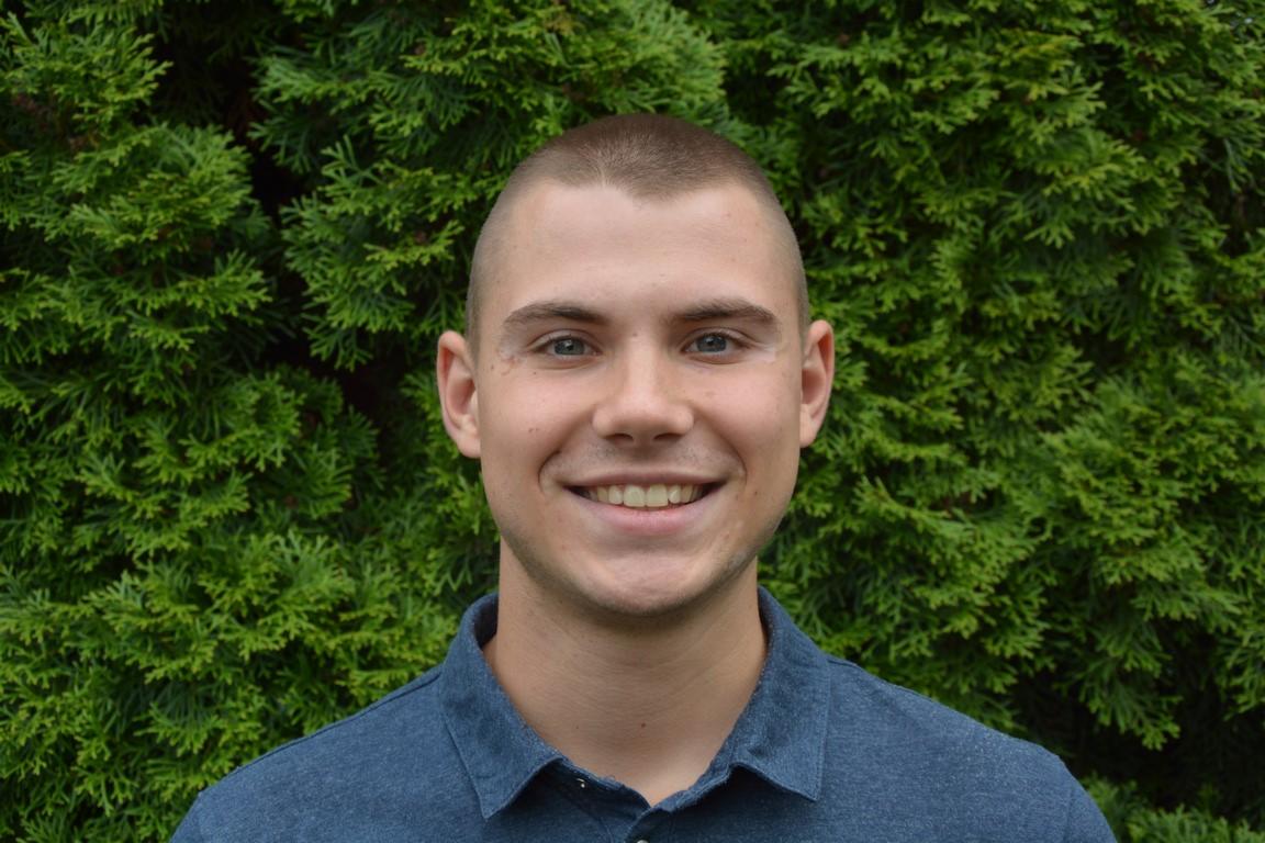 Uśmiechnięty mężczyzna w niebieskiej koszuli, z krótkimi włosami, na tle zielonego, gęstego żywopłotu. Portret.