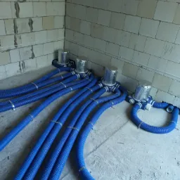 Niebieskie, karbowane przewody wentylacyjne rozchodzące się od czterech srebrnych, zaizolowanych anemostatów na surowym, betonowym podłożu w pomieszczeniu z białymi, nieotynkowanymi ścianami...