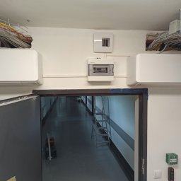 Dawid Sadło - Dwa białe klimatyzatory Fujitsu zamontowane pod sufitem w korytarzu biurowym, widoczna instalacja elektryczna w korytkach i skrzynka bezpiecznikowa, drabina w tle.