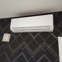 Biały klimatyzator ścienny zamontowany na ścianie z ciemną tapetą w romby, obok lampki nocnej i framugi drzwi.