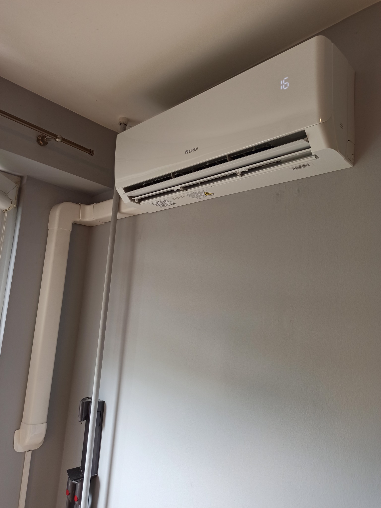 Biały klimatyzator Gree zamontowany na szarej ścianie w pomieszczeniu, widoczne rury instalacyjne w rogu, temperatura wyświetlana na urządzeniu to 16 stopni.
