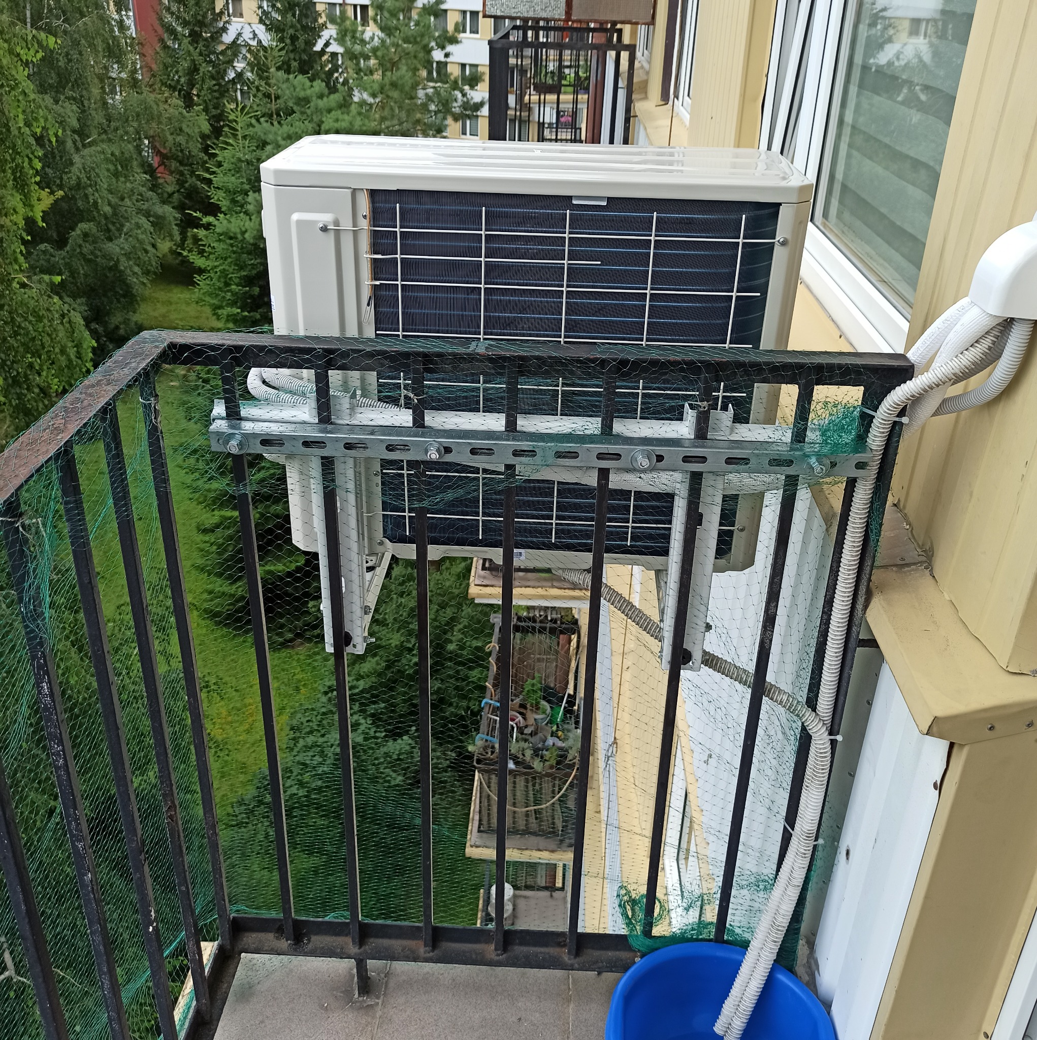 Zewnętrzna jednostka klimatyzatora zamontowana na balkonie bloku mieszkalnego, widoczna z góry przez metalową balustradę z siatką zabezpieczającą.