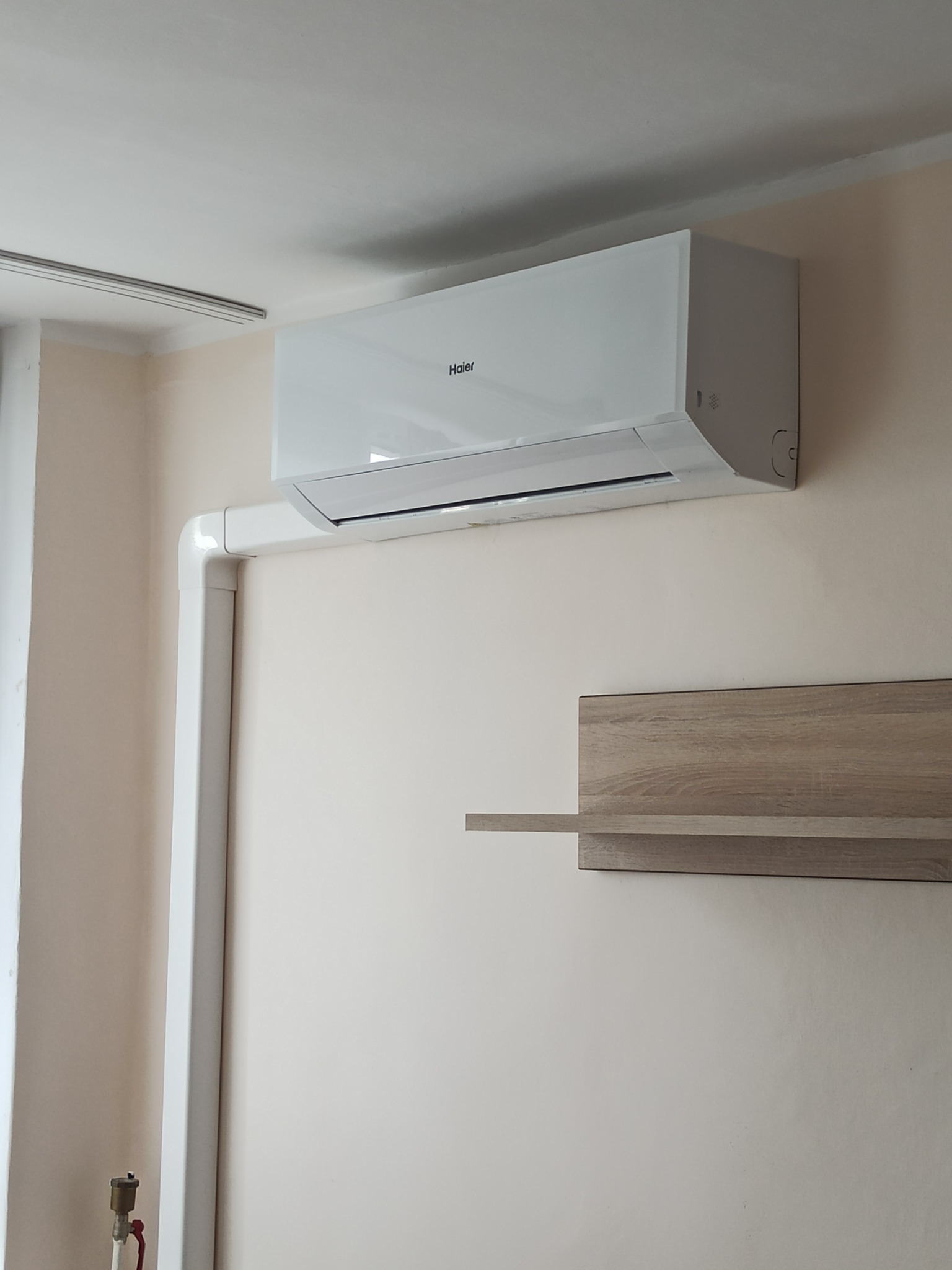 Biały klimatyzator Haier zamontowany na jasnej ścianie w pokoju, widoczne białe rury instalacyjne i półka z jasnego drewna.