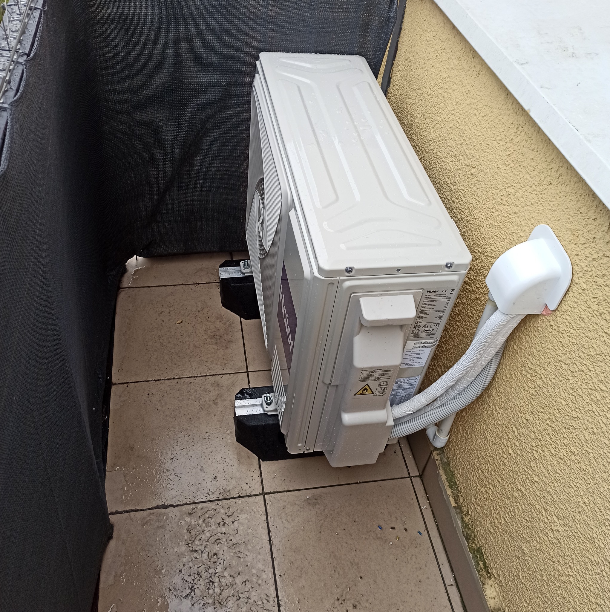 Zewnętrzna jednostka klimatyzatora Haier zamontowana na balkonie, widoczne przewody prowadzące do ściany budynku, czarne podkładki antywibracyjne pod urządzeniem.
