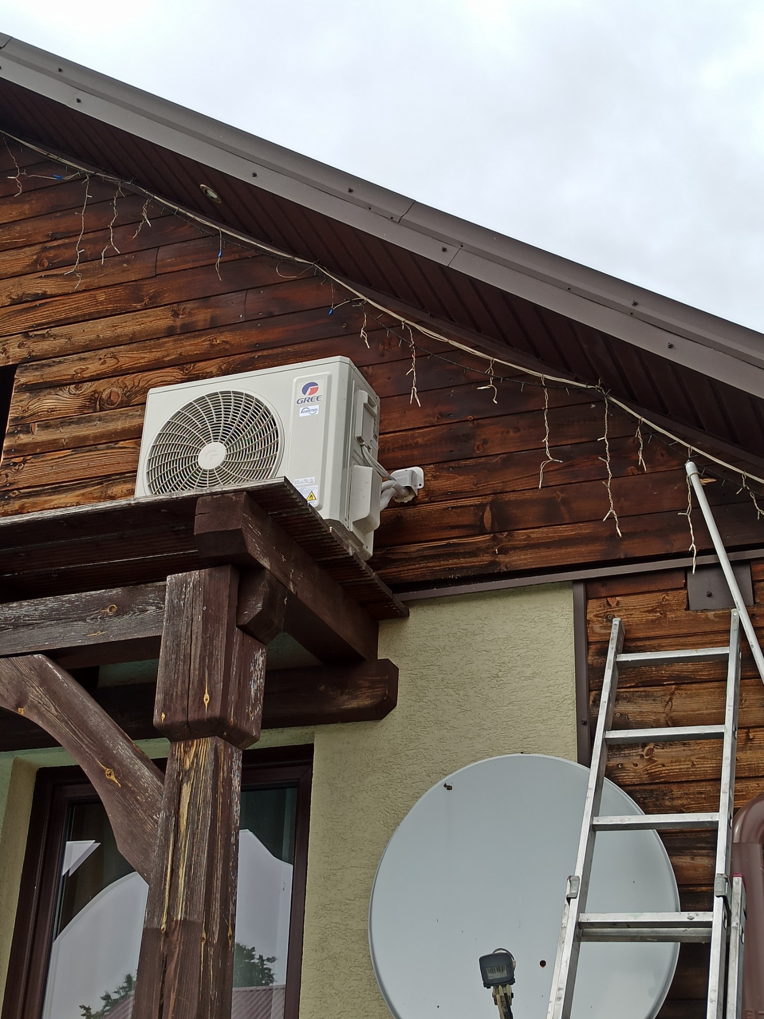 Zewnętrzna jednostka klimatyzatora marki Gree zamontowana na drewnianej konstrukcji przy ścianie domu z ociepleniem, obok przystawiona aluminiowa drabina, na ścianie wisi łańcuch lampek.
