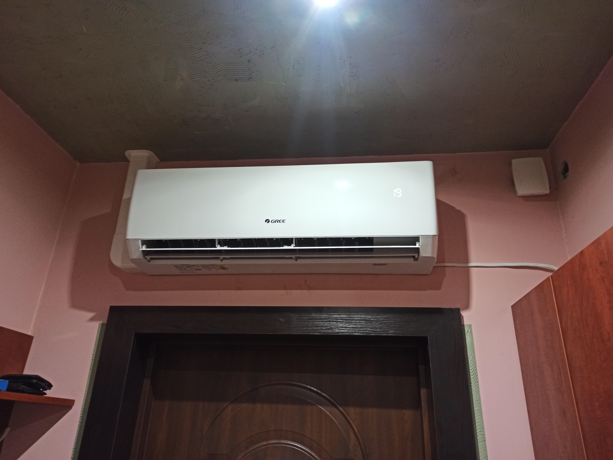 Biały klimatyzator marki Gree zamontowany nad ciemnym futryną drzwiową w pomieszczeniu o różowych ścianach i surowym, szarym suficie. Na wyświetlaczu widoczna temperatura 19 stopni.