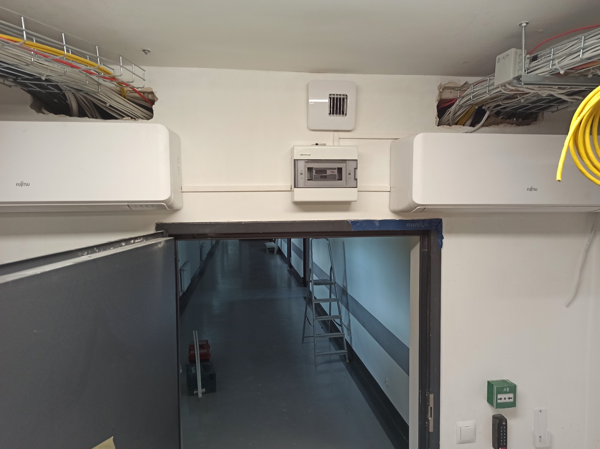 Dwa białe klimatyzatory Fujitsu zamontowane pod sufitem w korytarzu biurowym, widoczna instalacja elektryczna w korytkach i skrzynka bezpiecznikowa, drabina w tle.