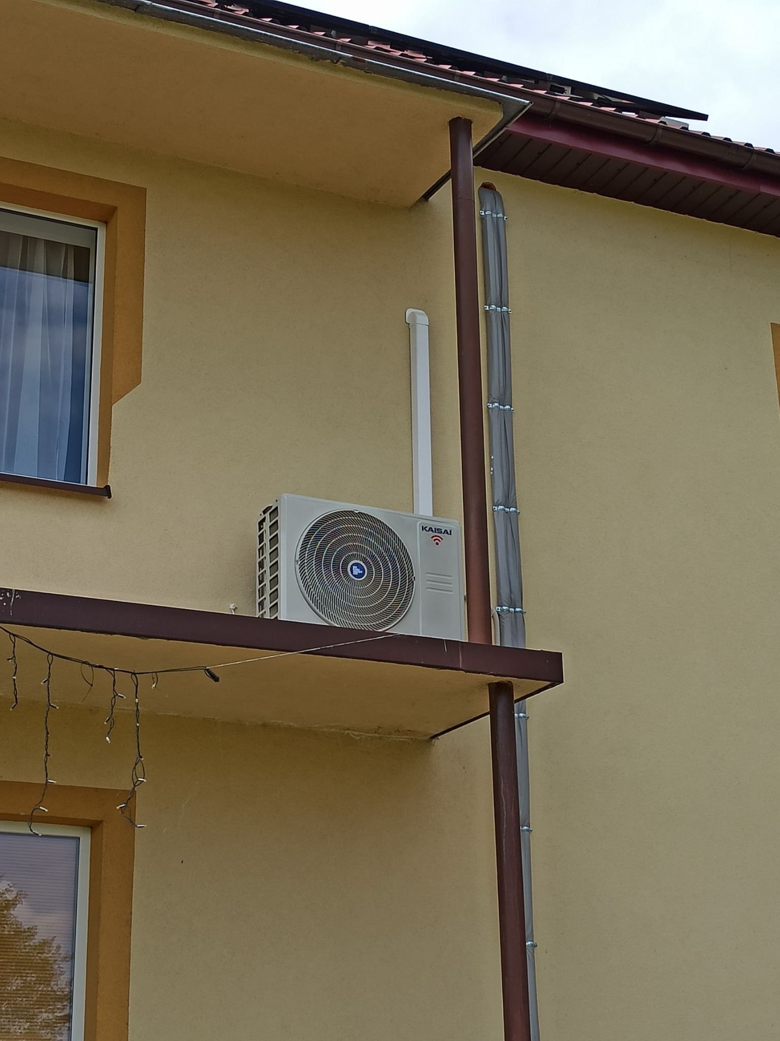 Zewnętrzna jednostka klimatyzacji Kaisai zamontowana na wsporniku przy ścianie budynku o beżowej elewacji, z widocznymi przewodami zabezpieczonymi osłonami.