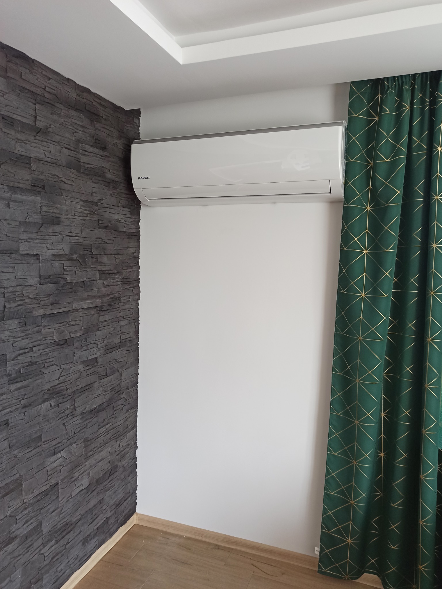 Biała jednostka klimatyzacyjna marki Kaisai zamontowana na białej ścianie, obok ściany z szarej imitacji kamienia i zielonych zasłon w złote geometryczne wzory.