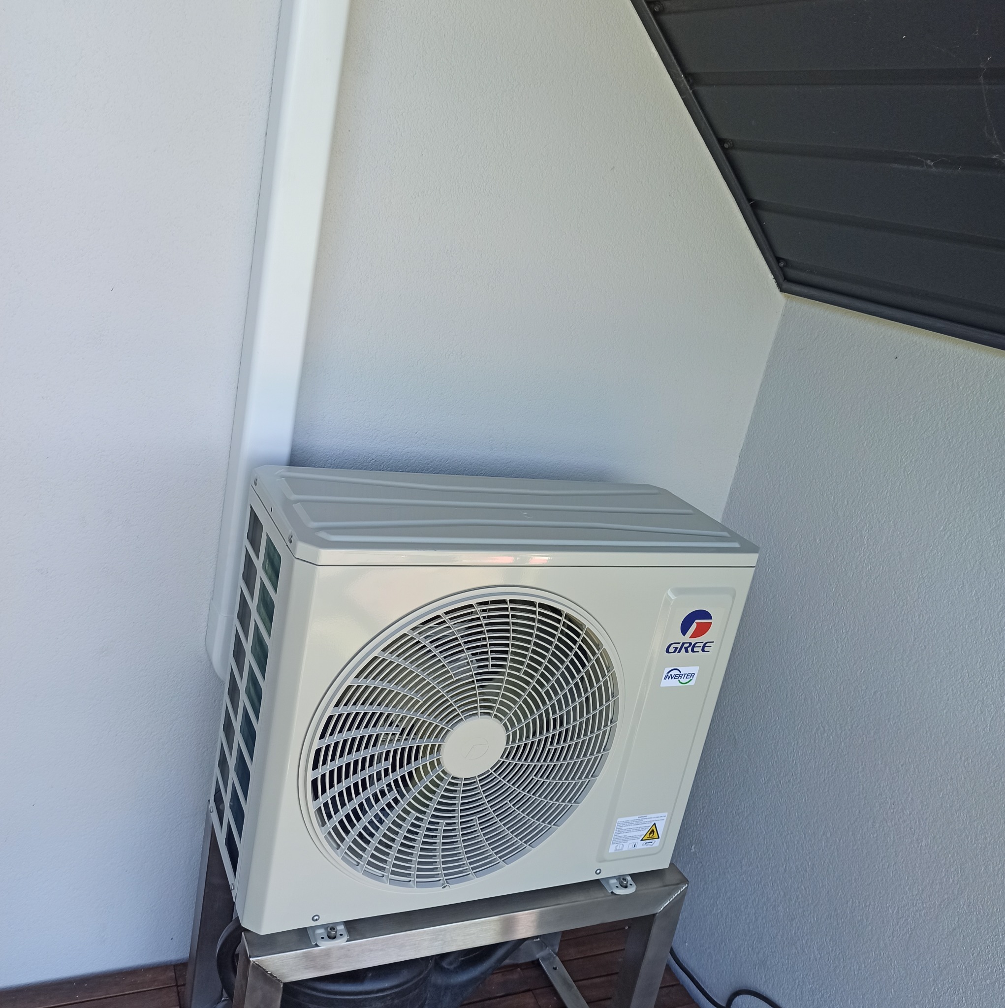 Zewnętrzna jednostka klimatyzatora marki Gree zamontowana na metalowym stelażu przy jasnej ścianie budynku, z widoczną rurą odprowadzającą skropliny i fragmentem dachu w rogu.