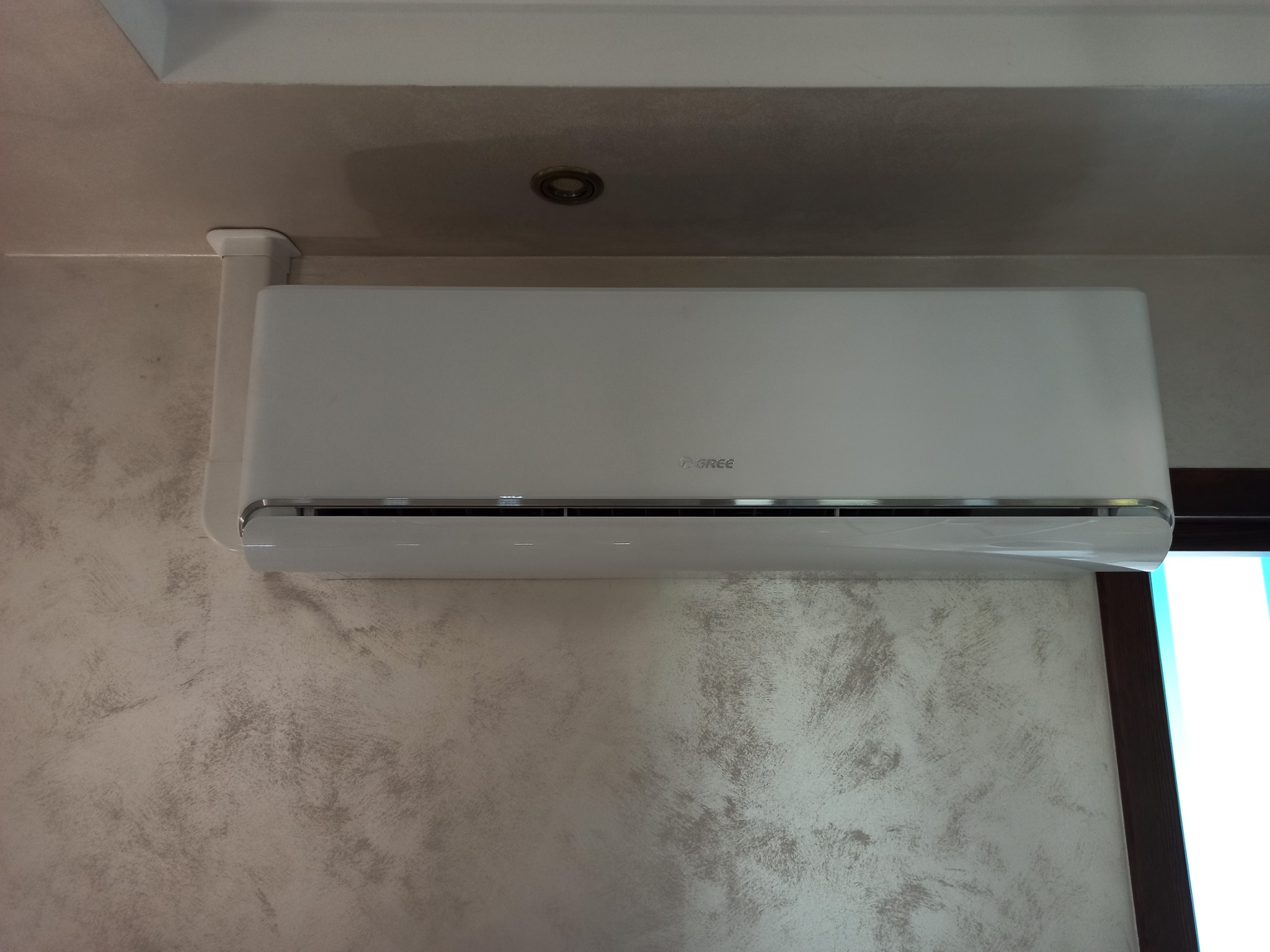 Biały klimatyzator marki Gree zamontowany na ścianie o fakturze betonu, widoczny fragment sufitu z oświetleniem punktowym.