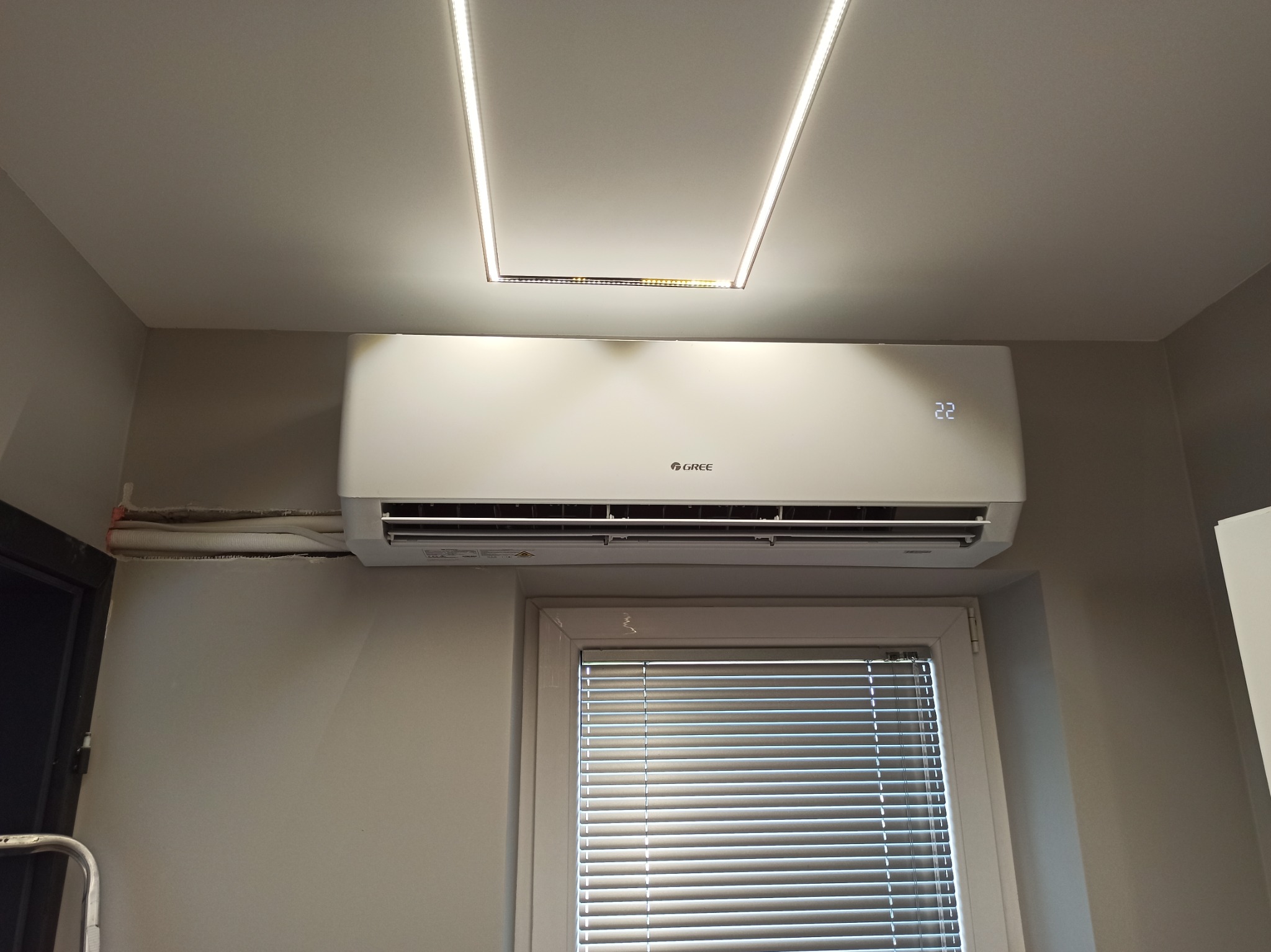Biała jednostka klimatyzacji marki Gree zamontowana na ścianie nad oknem z żaluzjami, widoczne rury instalacyjne przy suficie i oświetlenie LED w suficie podwieszanym.