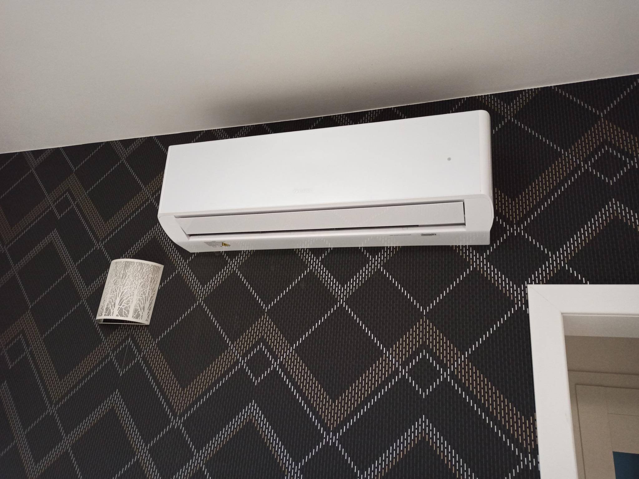Biały klimatyzator ścienny zamontowany na ścianie z ciemną tapetą w romby, obok lampki nocnej i framugi drzwi.