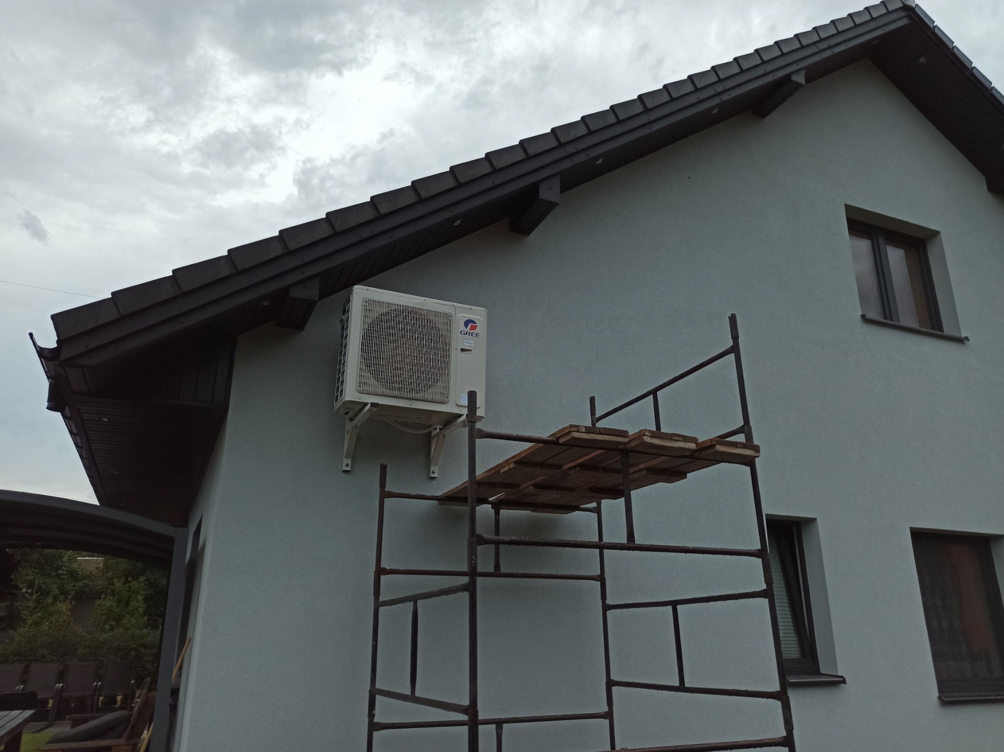 Biały klimatyzator marki Gree zamontowany na elewacji domu jednorodzinnego, widoczne rusztowanie z drewnianym podestem, pochmurne niebo w tle.