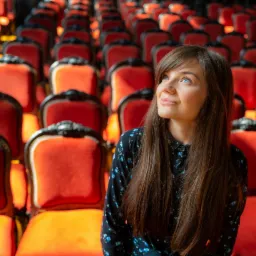 Portret kobiety z długimi brązowymi włosami na tle rzędów czerwonych krzeseł w teatrze, spoglądającej w górę w jasnym świetle.