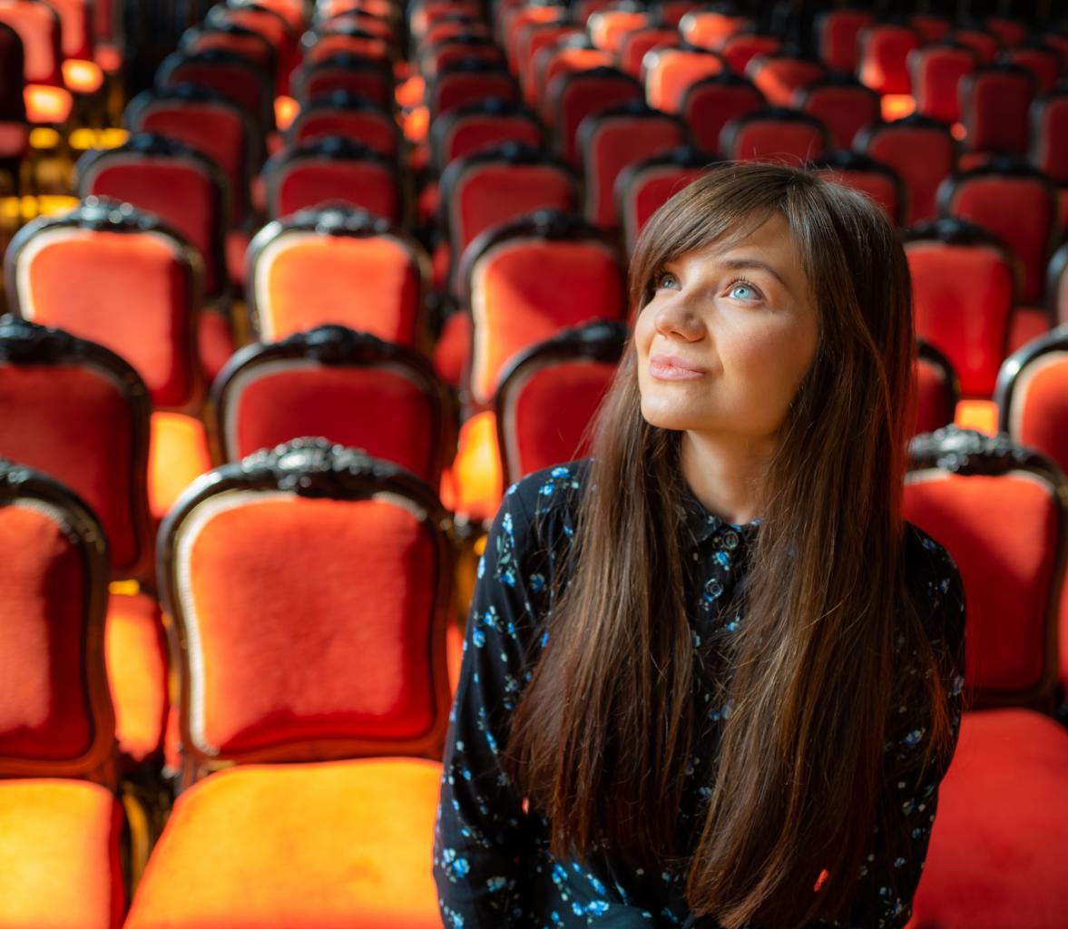 Portret kobiety z długimi brązowymi włosami na tle rzędów czerwonych krzeseł w teatrze, spoglądającej w górę w jasnym świetle.