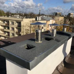 Widok z góry na komin z trzema metalowymi wkładami kominowymi z daszkami, na dachu pokrytym papą, z widocznymi budynkami mieszkalnymi w tle, w słoneczny dzień.