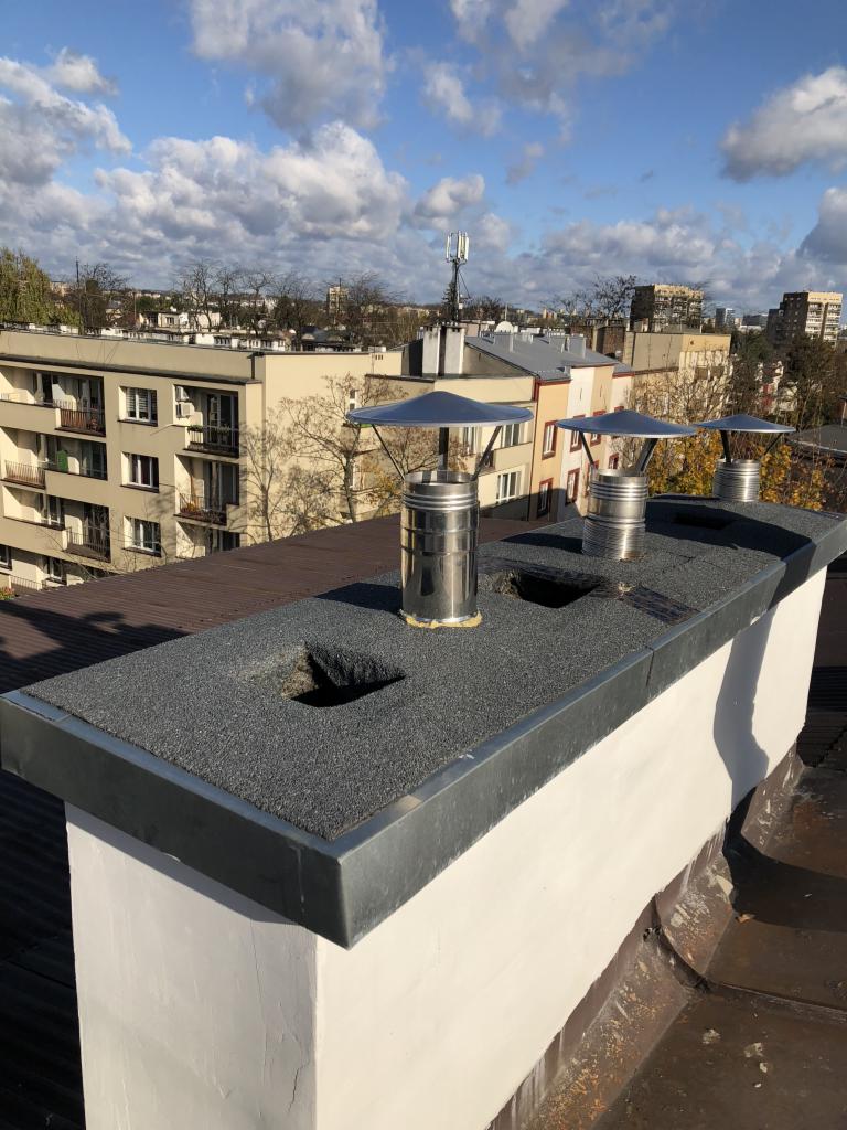 Widok z góry na komin z trzema metalowymi wkładami kominowymi z daszkami, na dachu pokrytym papą, z widocznymi budynkami mieszkalnymi w tle, w słoneczny dzień.