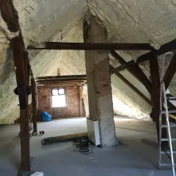 Poddasze z drewnianą konstrukcją dachu ocieplone pianką PUR, widoczny fragment muru z cegły, drabina i sprzęt budowlany.