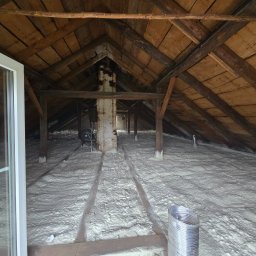 Izolacje Natryskowe Kielczyk Rafał Rafpur - Poddasze po izolacji pianką PUR w Trzciance. Widoczne drewniane elementy konstrukcji dachu i biała, równomierna warstwa izolacji. Metalowy przewód wentylacyjny w rogu.