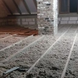 Izolacje Natryskowe Kielczyk Rafał Rafpur - Sypka izolacja termiczna wypełniająca przestrzenie między drewnianymi legarami na poddaszu, z ceglanym filarem i oknem w tle.