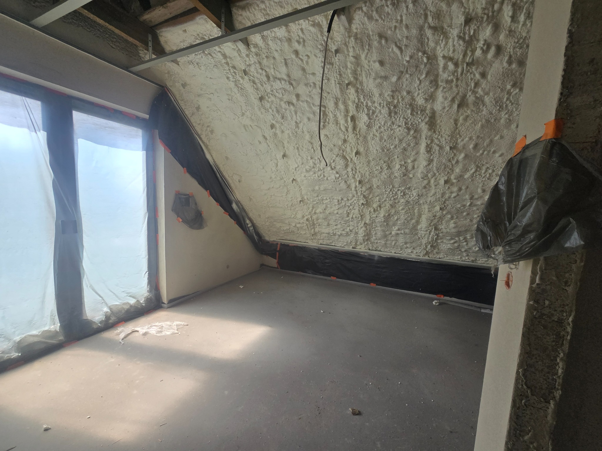 Poddasze po ociepleniu pianką PUR: widoczne białe, nierówne struktury pianki na skośnych ścianach i suficie, zabezpieczone folią okno, szary betonowy podkład.