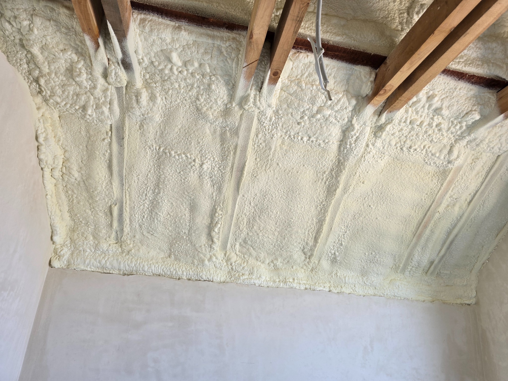 Poddasze z drewnianą konstrukcją i białą, natryskową izolacją piankową, widok z dołu, surowe ściany w tle.