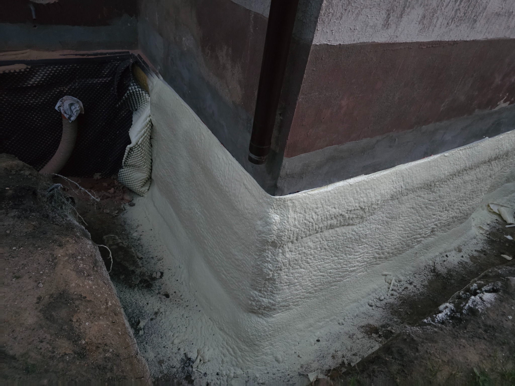 Szczegółowe ujęcie izolacji fundamentu pianką PUR, z widoczną strukturą pianki i elementami gruntu. Rura spustowa w tle.