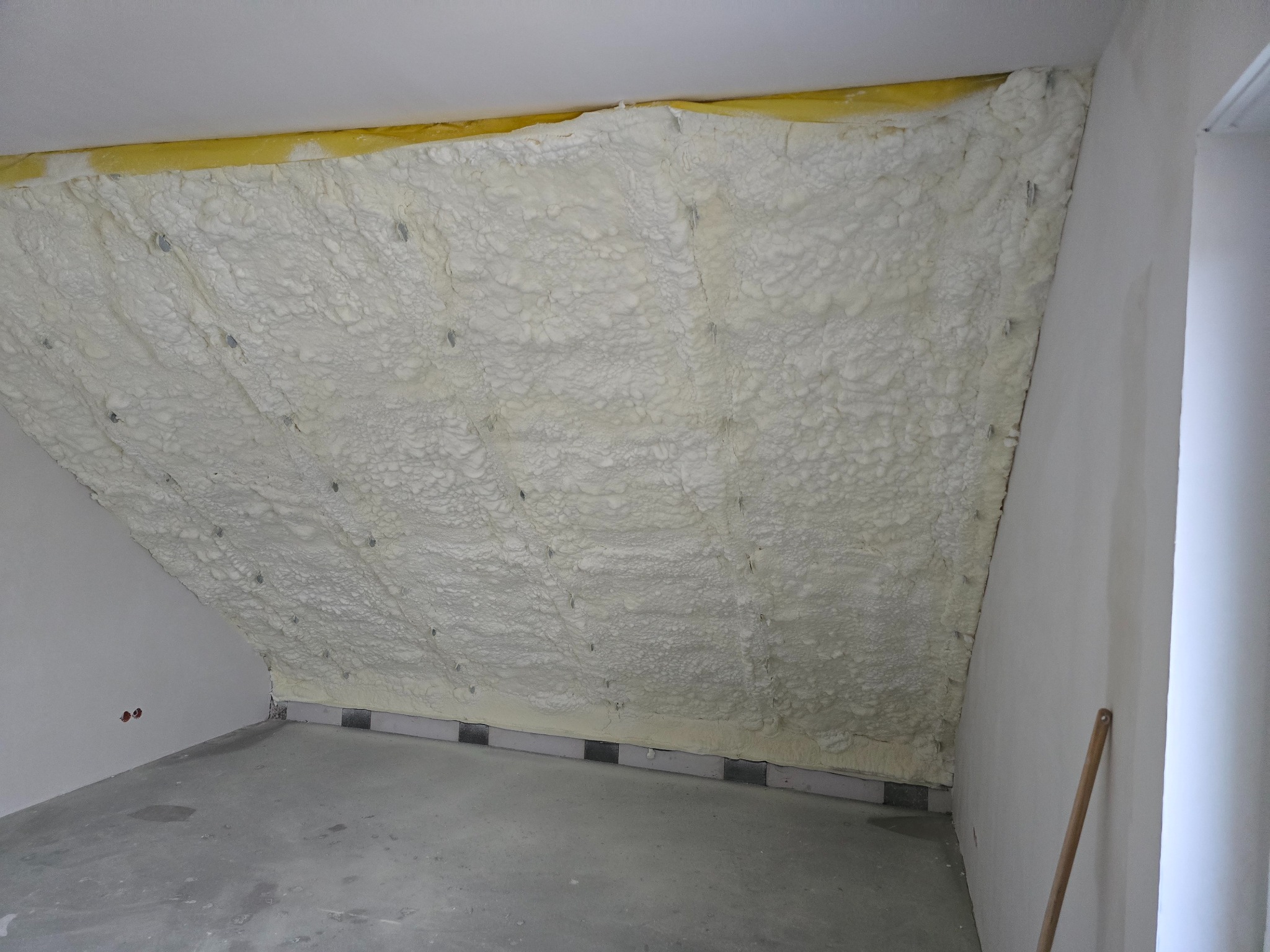 Poddasze po ociepleniu pianką PUR, widoczne mocowania, żółta warstwa izolacyjna przy suficie, surowy beton na podłodze, ściany przygotowane do wykończenia.