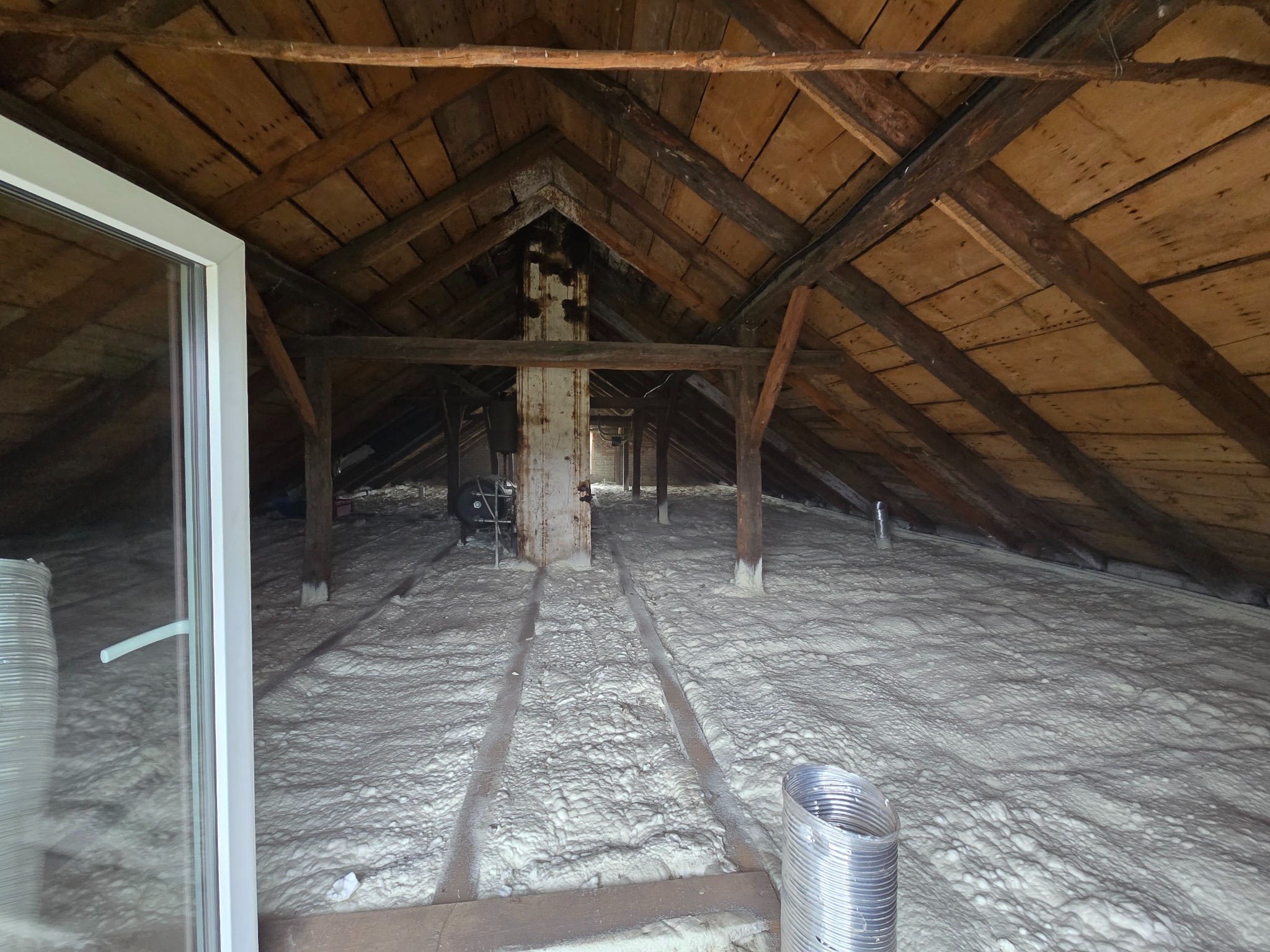 Poddasze po izolacji pianką PUR w Trzciance. Widoczne drewniane elementy konstrukcji dachu i biała, równomierna warstwa izolacji. Metalowy przewód wentylacyjny w rogu.