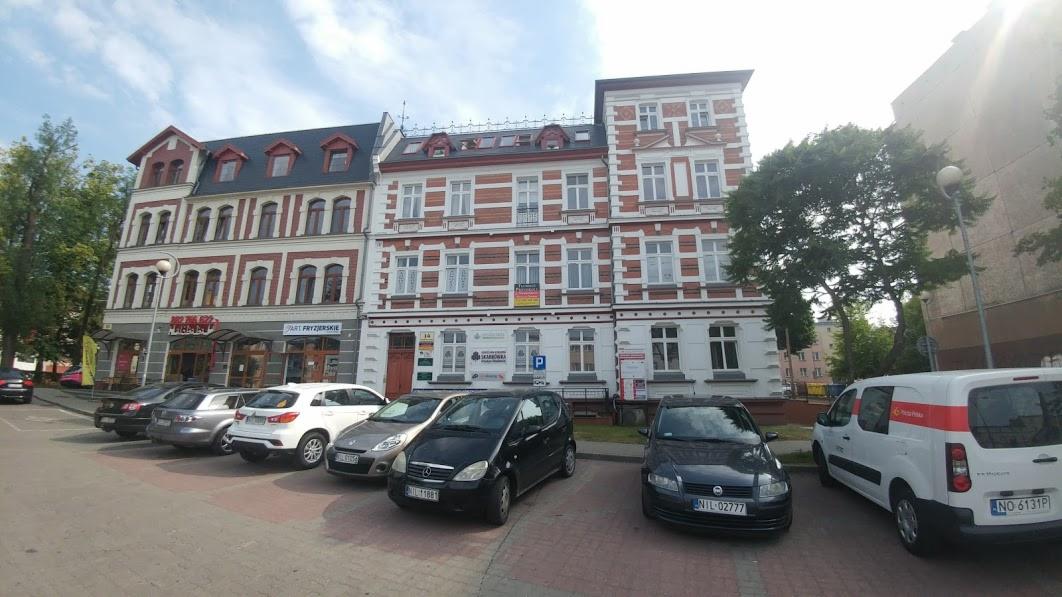 Elewacja kamienicy z biurami i zaparkowanymi samochodami w słoneczny dzień.