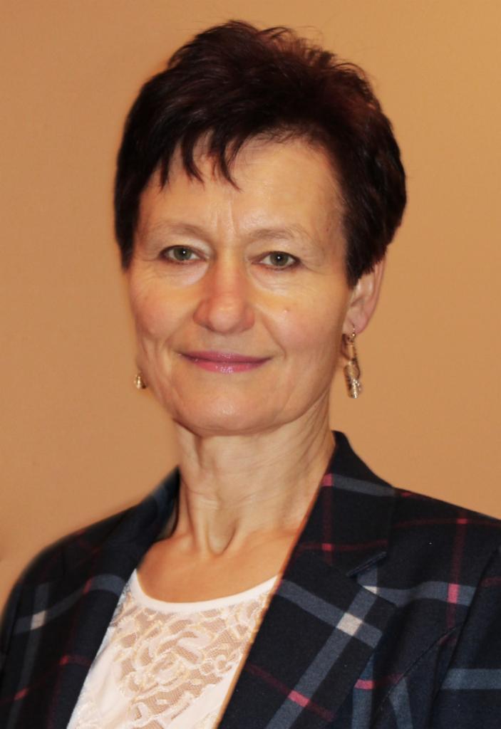 Portret uśmiechniętej kobiety w marynarce w kratę i koronkowej bluzce, z kolczykami, na jednolitym tle. Ciepłe oświetlenie.