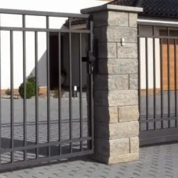 Nowoczesna, przesuwna brama metalowa w kolorze grafitowym, osadzona na słupkach z szarej kostki. Widoczny fragment ogrodzenia i garaż w tle.